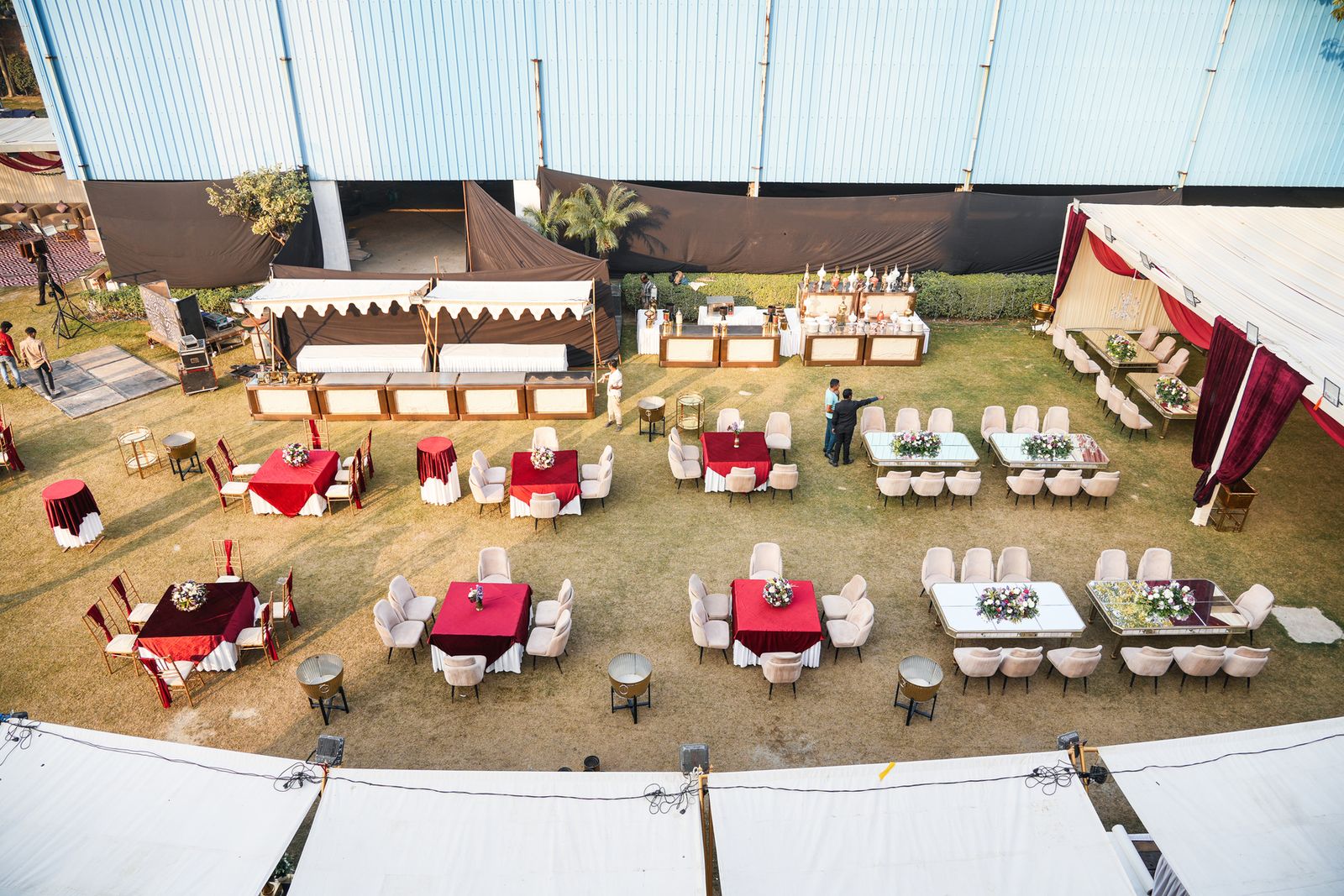Aerial view of Aarna Farms Noida lawn set for a daytime wedding — round dining tables with red velvet linens, cream chairs, canopy tents and live buffet counters