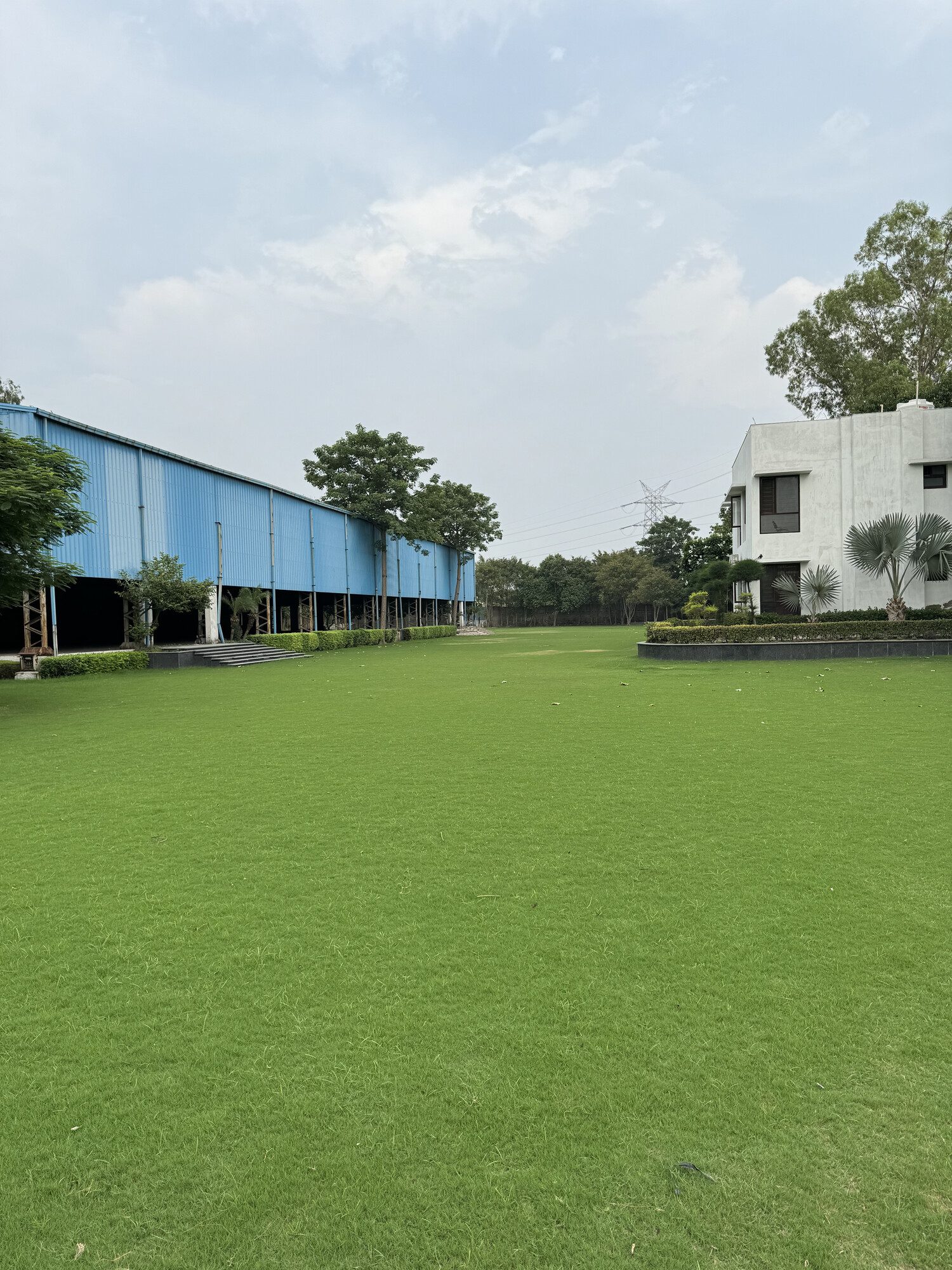Sprawling open green wedding lawn at Aarna Farms Sector-133 Noida — manicured grass stretching between the covered pavilion and the white farmhouse, a blank canvas venue for weddings