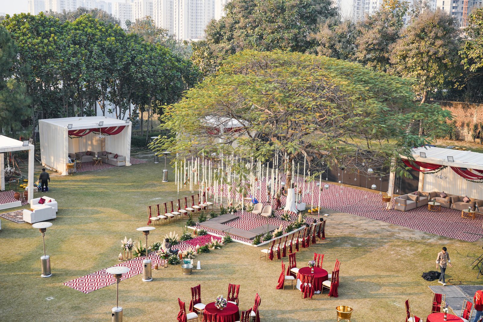 Outdoor wedding ceremony seating under a majestic tree at Aarna Farms Noida — pampas grass aisle, red and cream chairs, lounge cabanas and Noida city skyline in the background